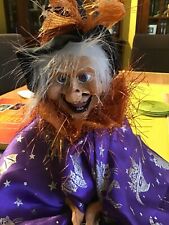 Hexenpuppe auf Besen reitend . Mit lilafarbenem Umhang und Hexenhut . 